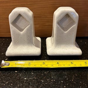 May include: Two speckled, off-white ceramic towel bar holders. Each has a rectangular base and a diamond-shaped cutout. The tape measure shows the holders are approximately 2 inches (5 cm) wide.