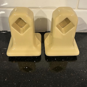 May include: Two matching, cream-colored ceramic towel holders. Each has a rectangular base and a diamond-shaped cutout in the center. The holders are likely for a bathroom or kitchen.