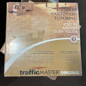 May include: A square package of TrafficMaster solid hardwood parquet flooring. The packaging features a light brown wood grain pattern, a 15-year residential warranty seal, and text detailing 10 square feet of coverage and installation features.