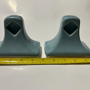 May include: Two matching teal-colored ceramic towel holders. Each holder has a square base, a curved body, and a diamond-shaped opening. The holders are approximately 4 inches wide, and 4 inches tall.