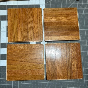 May include: Four square wooden floor samples with a medium brown oak grain pattern. The samples are arranged on a gray cutting mat with a grid pattern, showcasing the wood's natural texture and color variations.