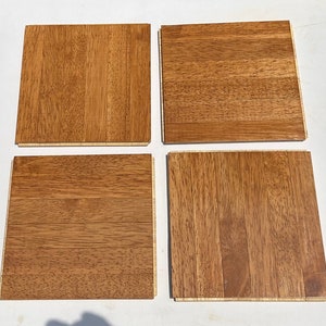 May include: Four square wooden flooring planks arranged in a grid pattern. Each plank features a natural medium brown wood grain finish with visible vertical and horizontal textures, showcasing the organic patterns and light tan edges of the wood.