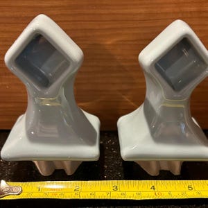 May include: Two light blue ceramic wall hooks. Each hook has a square top with a recessed area and a curved base. The hooks are displayed on a dark surface with a wooden background.