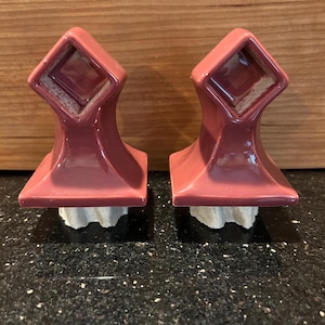 May include: Two matching maroon ceramic bottle stoppers. Each stopper has a square top with a diamond-shaped indentation and a flared base. The base is attached to a light-colored, textured cork.