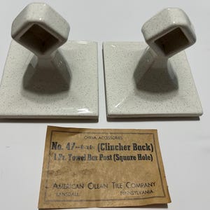 May include: Two vintage ceramic towel bar posts with a speckled off-white finish. Each post has a square base and a tilted, square-shaped top. A paper label with text is visible below the posts.