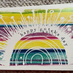 May include: A white card with a rainbow design and the text "happy gotcha day" printed in white.