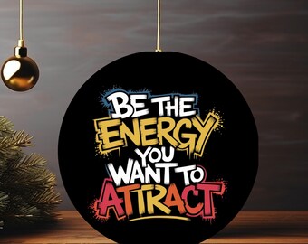 May include: A black round ornament with the text "BE THE ENERGY YOU WANT TO ATTRACT" in white, yellow, orange, and red. The ornament is hanging from a gold string. A gold ornament and a small Christmas tree are in the background.