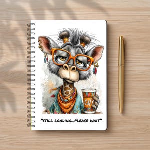 May include: A spiral-bound notebook featuring a graphic of a giraffe wearing orange glasses, a patterned scarf, and earrings, holding a cup labeled "Cafe". The text "STILL LOADING...PLEASE WAIT" is printed below. A gold pen lies beside the notebook.