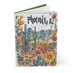 May include: A hardcover journal with a green spine and a colorful illustration of Phoenix, AZ. The cover features the city's name, cacti, flowers, and a desert landscape with mountains in the background.
