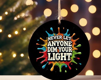 May include: Black round ornament with the text "NEVER LET ANYONE DIM YOUR LIGHT" in white, orange, and yellow. The text is surrounded by colorful paint splatters. A natural twine loop is at the top for hanging.