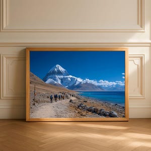 May include: A framed landscape photograph of a mountain range with a snow-capped peak, a blue lake, and a path with people walking. The sky is a clear, bright blue. The frame is made of light-colored wood.