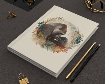 Puede incluir: Un diario de color crema con una ilustración de acuarela de dos elefantes abrazándose dentro de una corona floral. El diario tiene lomo y páginas blancas. También se ven lápices dorados y negros y clips dorados.