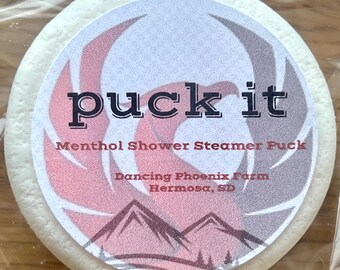 Può includere: Un disco doccia al mentolo bianco e rotondo con la scritta "puck it" in nero. L'etichetta presenta un design rosso e grigio con il testo "Menthol Shower Steamer Puck" e "Dancing Phoenix Farm Hermosa, SD".