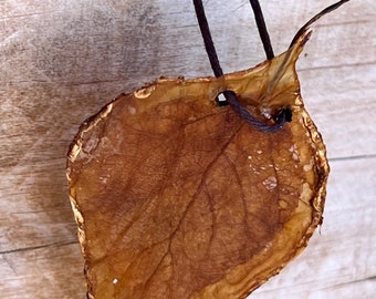 Può includere: Un ciondolo a forma di foglia conservata, di colore marrone con bordi dorati, appeso a un cordino marrone scuro. La foglia mostra venature dettagliate, risaltata su uno sfondo di legno.