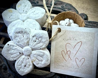 Può includere: Bagnoschiuma bianchi a forma di fiore con macchie scure sono disposti con un piccolo guscio di cocco pieno di bagnoschiuma a forma di cuore. È attaccato un'etichetta con "Grazie" e tre cuori.