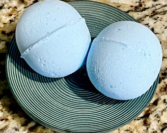 Può includere: Due bombe da bagno azzurre con superficie strutturata, appoggiate su un piatto verde scuro con un design a cerchi concentrici. Le bombe da bagno sono sferiche con una cucitura visibile. Il piatto poggia su una superficie maculata marrone chiaro.