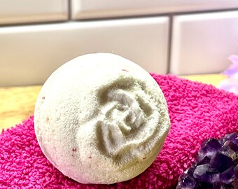 Può includere: Una bomba da bagno rotonda, bianco sporco, con un disegno a rosa, appoggiata su un asciugamano rosa acceso. La bomba da bagno ha una superficie strutturata e piccole macchie rosse. Un gruppo di cristalli viola è visibile sullo sfondo.