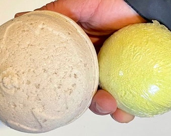 Può includere: Due bombe da bagno sferiche. Una è di colore beige chiaro con una superficie strutturata. L'altra è di colore giallo brillante ed è avvolta in plastica trasparente. Le bombe da bagno sono tenute in mano da una persona.