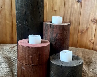 Può includere: Una collezione di portacandele rustici in legno di varie altezze e colori. Il più alto è scuro, mentre gli altri sono rossi e marroni. Ognuno contiene una candela bianca, creando un'atmosfera calda e accogliente. La venatura del legno è visibile.