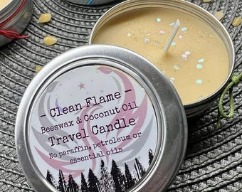 Può includere: Primo piano di una candela da viaggio in una latta d'argento. La candela è di colore giallo chiaro con uno stoppino bianco e glitter. L'etichetta recita "Clean Flame Travel Candle" ed elenca cera d'api e olio di cocco come ingredienti. Altre candele sono visibili.