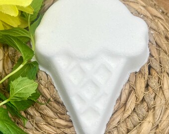 Può includere: Una bomba da bagno bianca a forma di gelato con un motivo a cono waffle. La bomba da bagno è appoggiata su un tappetino circolare intrecciato, con foglie verdi e petali di fiori gialli sullo sfondo.