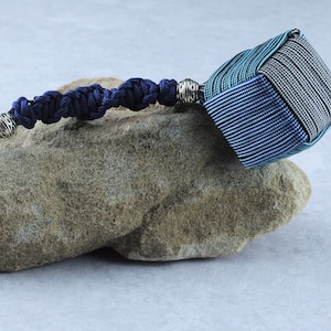 May include: A blue and teal braided key chain with a silver metal clasp and a silver bead. The key chain is tied into a square shape at the end.