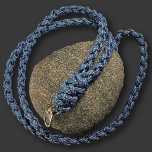 May include: A blue and black braided lanyard with a silver-colored clasp. The lanyard is draped over a gray, textured stone against a dark gray background. The lanyard is made of a woven material.