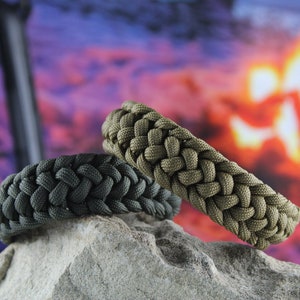 May include: Two braided paracord bracelets in dark green and olive green, resting on a gray rock. The bracelets are made with a woven pattern and are a popular accessory for outdoor enthusiasts.