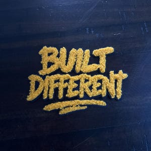May include: A yellow embroidered patch with the words "BUILT DIFFERENT" in a bold, textured font. The patch is set against a dark blue background, creating a striking contrast. The lettering has a fuzzy, three-dimensional appearance.