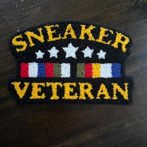 May include: Black, textured patch with the words "SNEAKER VETERAN" in yellow, arched across the top and bottom. Five white stars are centered above a multi-colored stripe design. The patch is likely for a sneaker enthusiast.