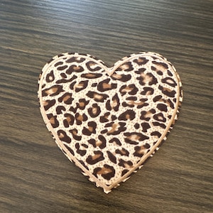 May include: A heart-shaped coaster with a leopard print design. The coaster features a tan background with brown spots and a tan border. The coaster is placed on a dark brown wooden surface.
