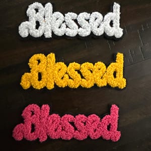 May include: Three embroidered patches with the word "Blessed" in a script font. The patches are white, yellow, and pink, each with a black border. The patches have a textured, fluffy appearance.