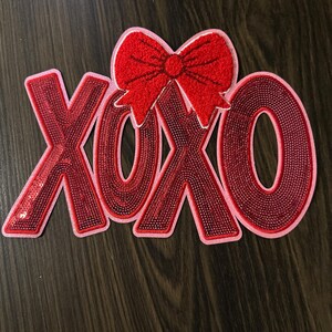 May include: A red sequined XOXO sign with a red bow on top. The letters are outlined in pink and filled with red sequins. The bow is made of red felt. The sign is on a dark wood surface.