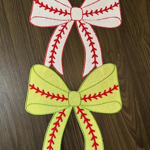 May include: Two decorative fabric bows. The top bow is white with red stitching, and the bottom bow is light green with red stitching. Both bows have a baseball-like stitch pattern. The bows are on a dark wood-grain surface.