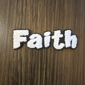 May include: A white chenille patch with a black border featuring the word Faith in a rounded, bubbly font. The letters have a textured, fuzzy surface and are connected to form a single piece, creating a bold, high-contrast design for fabric.