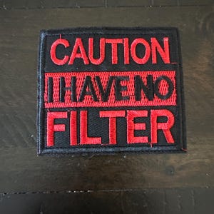 May include: A square embroidered patch with a black background and red border. The text CAUTION is embroidered in red at the top. The middle section features a red horizontal bar with the black text I HAVE NO in the center. The word FILTER is embroidered in red at the bottom.