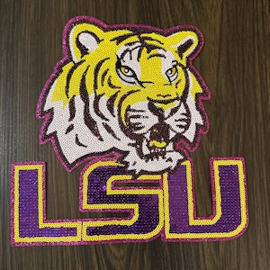 May include: A sequined patch featuring a tiger head and the letters "LSU". The tiger is yellow with brown stripes and a pink outline. The letters are purple with a yellow outline. The patch is likely for apparel or accessories.