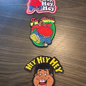 May include: Three embroidered patches featuring cartoon characters. The top patch shows a character in a red shirt with the text "Hey, Hey, Hey." The middle patch has a character labeled "Fat Gilbert." The bottom patch displays a character's face with "Hey Hey Hey."