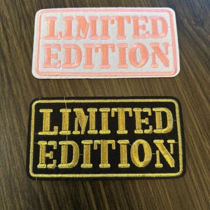 May include: Two rectangular patches with the text "LIMITED EDITION". One patch has a white background with pink lettering and border. The other patch has a black background with gold lettering and border. Both patches are embroidered.