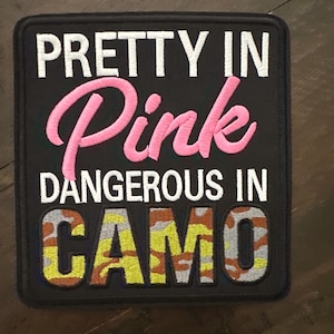 May include: A square embroidered patch with a black background. The text reads PRETTY IN in white, Pink in cursive pink thread, DANGEROUS IN in white, and CAMO in a camouflage pattern of green, brown, and grey thread with a black stitched border.