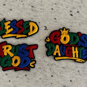May include: Three embroidered patches with black outlines and multi-colored lettering. Patch 1 reads BLESSED. Patch 2 reads TRUST GOD. Patch 3 reads GOD'S DAUGHTER with a small crown icon. The letters feature red, blue, green, and yellow colors.