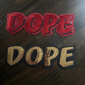 May include: Two sequined patches spelling the word "DOPE". One patch is red with black trim, the other is gold with black trim. The patches are likely for clothing or accessories.
