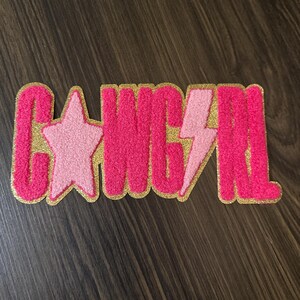 May include: A pink patch with the word "GANGSTER" in a bold, textured font. The letters are outlined in gold, with a pink star and lightning bolt incorporated into the design. The patch is on a dark wood surface.