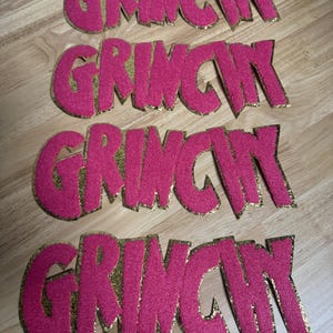 May include: Four pink felt cutouts spelling "GRINCH" with gold glitter edging. The letters are in a bold, stylized font. The cutouts are arranged on a wooden surface.
