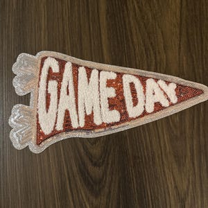 May include: A pennant-shaped patch with the words "GAME DAY" in white, textured lettering. The pennant has a red and orange border with a sequined silver edge. The background is a dark wood grain.