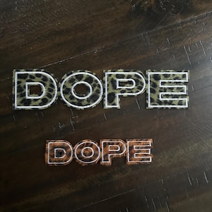 May include: Two patches spelling the word "DOPE". The top patch has a leopard print pattern with a white outline. The bottom patch has a brown and tan leopard print pattern with a white outline.