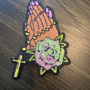 May include: Embroidered patch featuring praying hands with pink fingernails, a gold cross, and a sequined rose in shades of pink and green. The patch has a black glitter border and is set against a dark wood grain background.