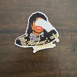 May include: An embroidered patch featuring a stylized figure with dark hair, wearing a black jacket with gold accents, and holding a silver-textured glove over their eyes. The name Michael Jackson is stitched in gold cursive script at the bottom.
