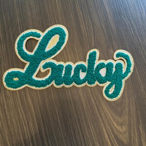 May include: A green and gold embroidered patch with the word "Lucky" in a script font. The word is in a textured green fabric with a gold border and a white outline. The patch is on a dark wood surface.