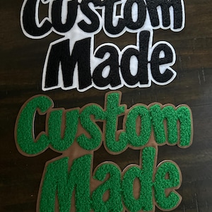 May include: Two embroidered patches with the words "Custom Made". The top patch has black letters with a white border. The bottom patch has green letters with a brown border. The patches are on a dark wood surface.